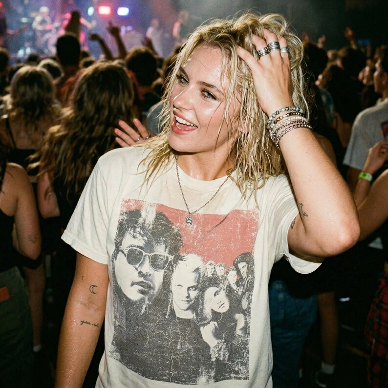 A woman with wet hair wearing a white t-shirt with a graphic of five people in a red box, smiling in a crowd at a concert.