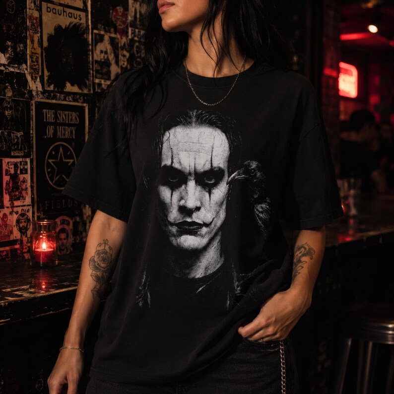 A person with tattoos on their arms wears a black t-shirt featuring a grayscale portrait of Brandon Lee as The Crow, with a crow perched on his shoulder.