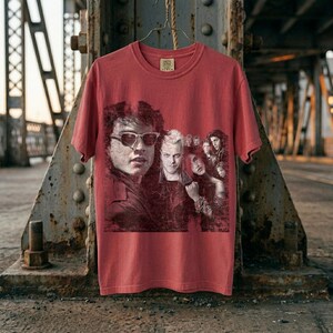 A reddish-brown t-shirt with a graphic of five people in a red box, hanging on a hanger against a rusty metal structure.