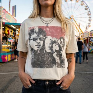 A person with blonde hair stands outdoors at a carnival, wearing a cream-colored t-shirt with a distressed graphic of five people, including two men wearing sunglasses.