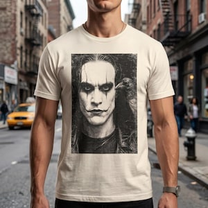 A man stands on a city street, wearing a light beige t-shirt with a grayscale portrait of Brandon Lee as The Crow, with a crow on his shoulder.