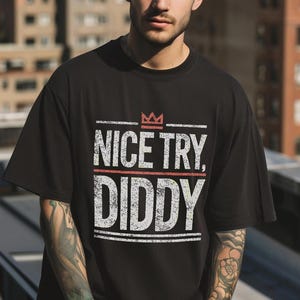 A person with tattoos on their arms wears a black t-shirt with "NICE TRY, DIDDY" in distressed white and red text, with a small red crown above the text.