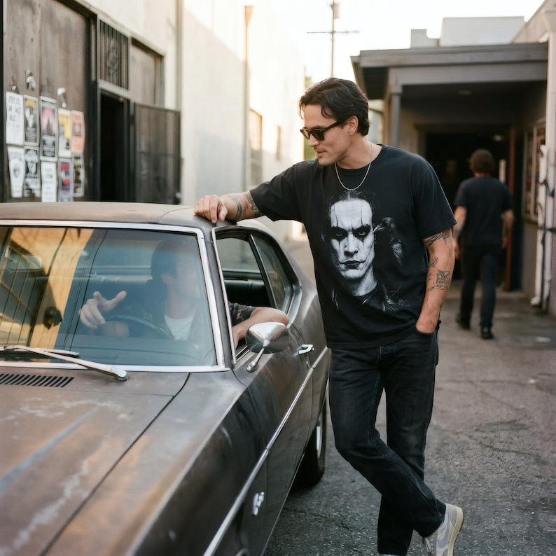 A man leans against a vintage car, wearing a black t-shirt with a grayscale portrait of Brandon Lee as The Crow.