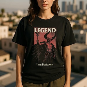 Puede incluir: Camiseta negra con un gráfico rojo y negro de un demonio con cuernos sosteniendo un bastón, con la palabra "LEGEND" arriba y "I am Darkness." abajo. La camiseta es de cuello redondo.
