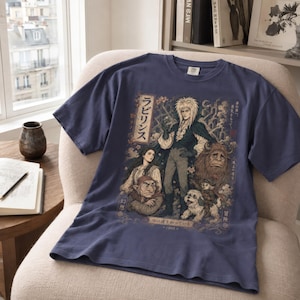 May include: A slate blue short-sleeve t-shirt featuring a detailed graphic print of characters from the film Labyrinth, including Jareth, Sarah, Hoggle, Ludo, and Sir Didymus, with Japanese text and cherry blossom accents integrated into the design.