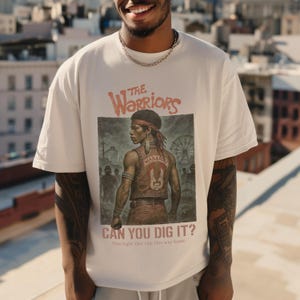 Könnte beinhalten: Ein weißes T-Shirt mit einem Grafikdruck eines Charakters aus dem Film "The Warriors". Die Grafik zeigt den Charakter in einer Lederjacke mit dem Wort "Warriors" auf dem Rücken. Der Text "Can You Dig It?" ist unter der Grafik gedruckt, zusammen mit dem Slogan "One night. One city. One way home."