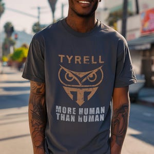 May include: Dark grey t-shirt featuring the word "TYRELL" above a brown owl design and the text "MORE HUMAN THAN HUMAN" below. The shirt has a crew neck.