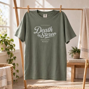The same muted green t-shirt with Death by Stereo text hanging on a wooden clothing rack.