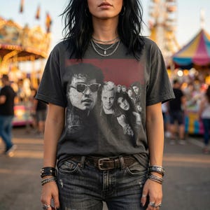 A person wearing a dark gray t-shirt with a graphic of five people in a red box, with a carnival in the background.