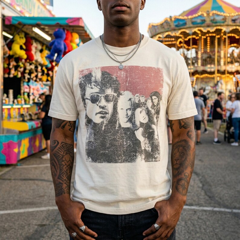 A person with dark skin and tattoos on their arms stands outdoors at a carnival, wearing a cream-colored t-shirt with a distressed graphic of five people, including two men wearing sunglasses.