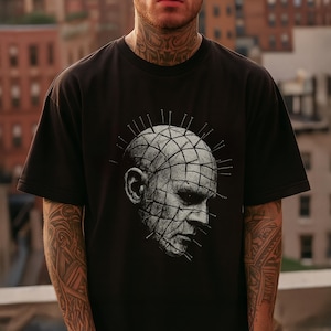 May include: Black t-shirt featuring a detailed grayscale graphic of a person's head with a grid pattern and nails. The design is centered on the front of the shirt. The shirt has short sleeves and a crew neck.