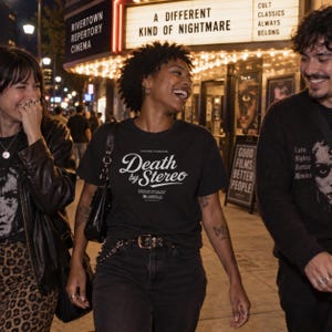 Three people smile and walk down a city street at night near a theater marquee, with the center person wearing a black Death by Stereo t-shirt.