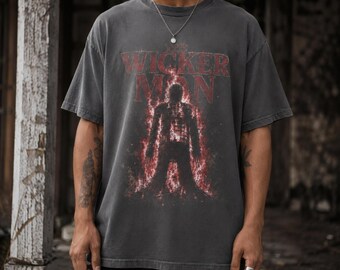 May include: A charcoal gray short-sleeve t-shirt featuring a graphic print of a figure engulfed in red flames. The text THE WICKER MAN is printed in a distressed red font above the central figure. The shirt has a crew neckline and a relaxed fit.