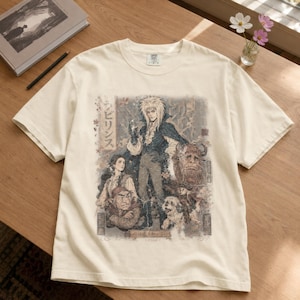 May include: A cream-colored short-sleeve t-shirt featuring a detailed graphic print of characters from the film Labyrinth. The design includes a central figure with spiky hair, a woman, a goblin, a large furry creature, and a small dog, with Japanese text.