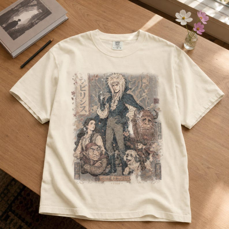 May include: A cream-colored short-sleeve t-shirt featuring a detailed graphic print of characters from the film Labyrinth. The design includes a central figure with spiky hair, a woman, a goblin, a large furry creature, and a small dog, with Japanese text.