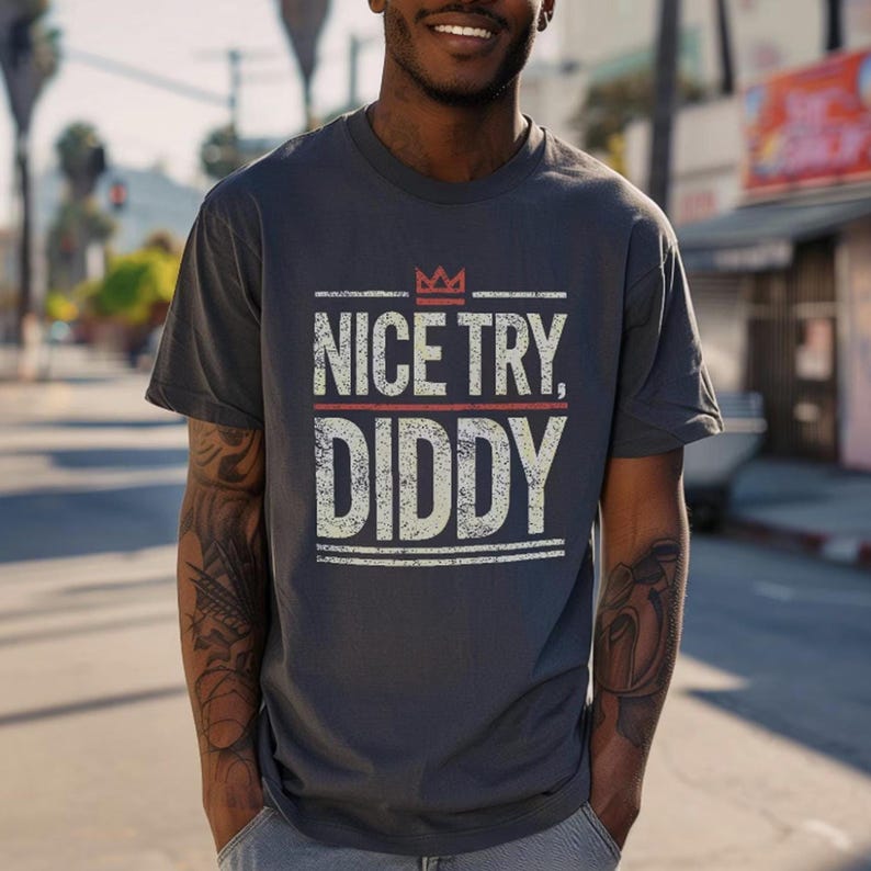 A person with short, dark hair and tattoos on their arms wears a dark gray t-shirt with "NICE TRY, DIDDY" in distressed white and red text, with a small red crown above the text.