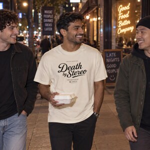 Three people walk on a city sidewalk at night, with the center person wearing an off-white t-shirt featuring black Death by Stereo text and holding a takeout container.
