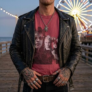 A person with tattoos on their hands and arms stands on a pier, wearing a black leather jacket and a dark red t-shirt with a distressed graphic of five people, including two men wearing sunglasses.
