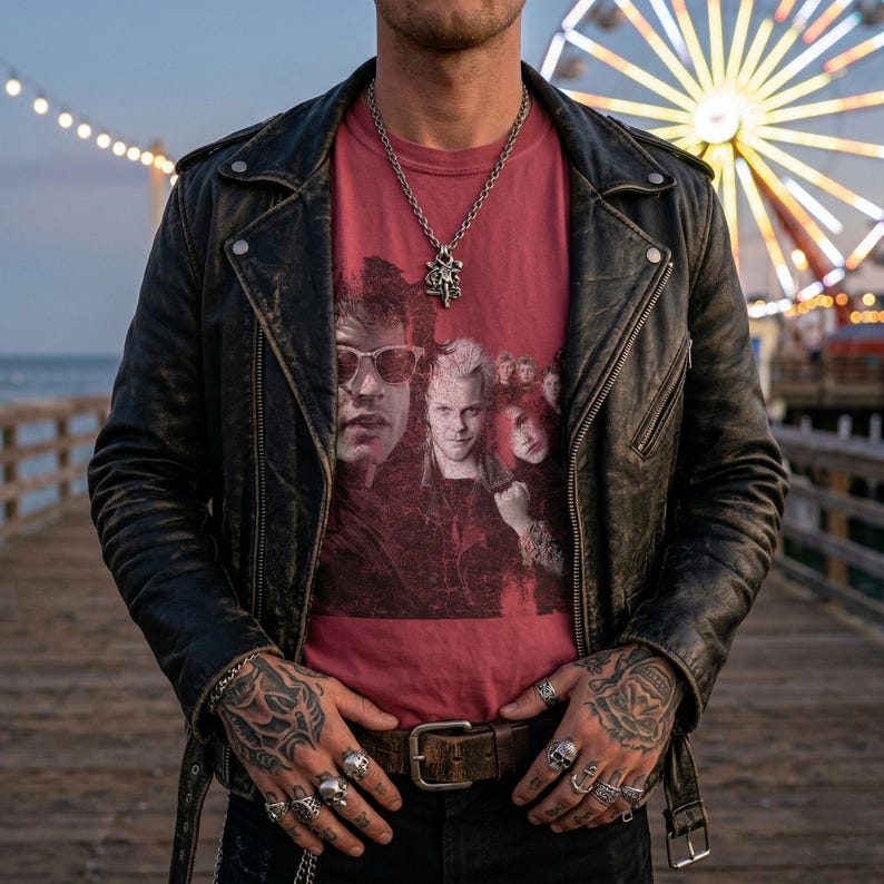 A person with tattoos on their hands and arms stands on a pier, wearing a black leather jacket and a dark red t-shirt with a distressed graphic of five people, including two men wearing sunglasses.