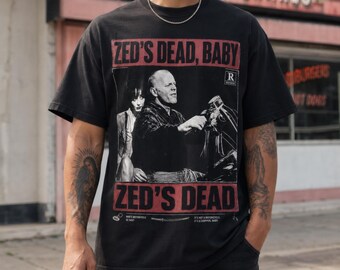 May include: A black short-sleeve t-shirt featuring a graphic print of a scene from the movie Pulp Fiction. The print includes the text ZED'S DEAD, BABY and ZED'S DEAD, along with a black and white image of two characters on a motorcycle.