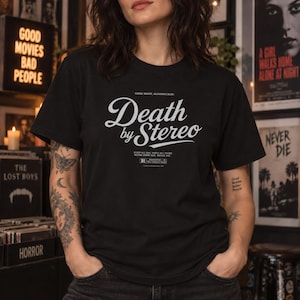 A person with tattoos wears a black t-shirt printed with the white text Death by Stereo in a record store setting.