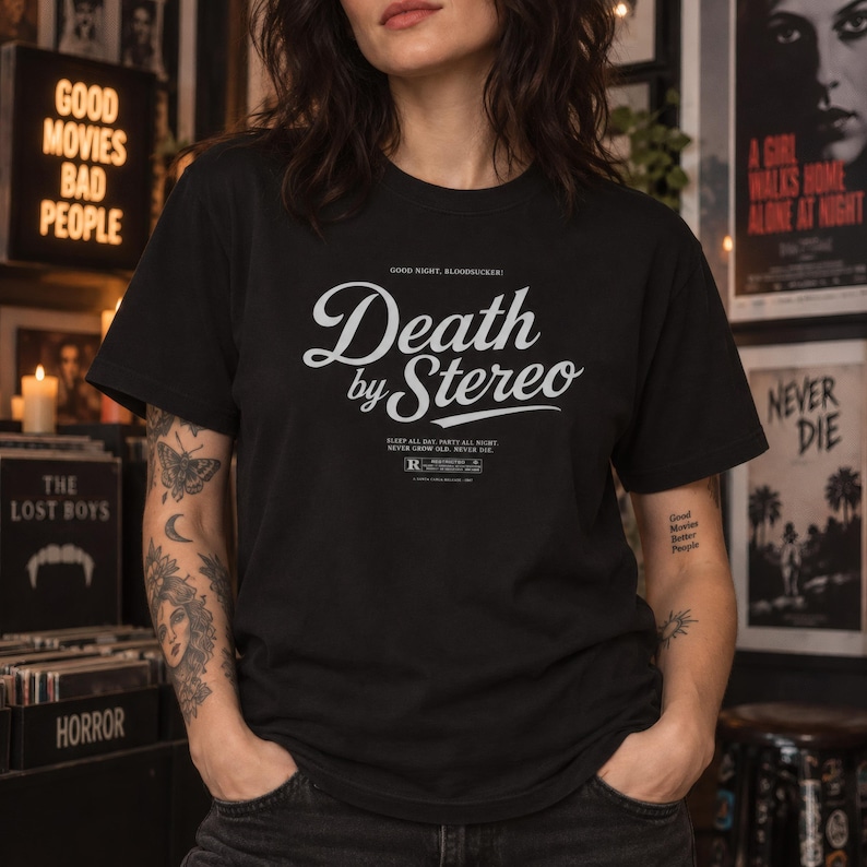 A person with tattoos wears a black t-shirt printed with the white text Death by Stereo in a record store setting.
