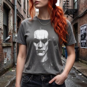 A person with red hair stands in an alley, wearing a dark gray t-shirt with a grayscale portrait of Brandon Lee as The Crow.