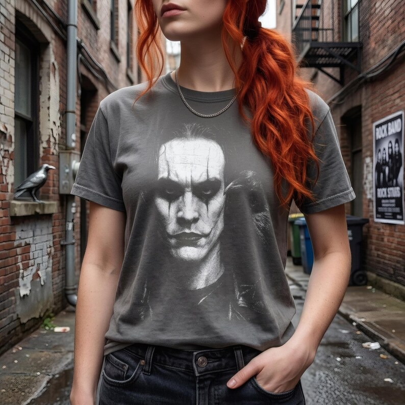 A person with red hair stands in an alley, wearing a dark gray t-shirt with a grayscale portrait of Brandon Lee as The Crow.