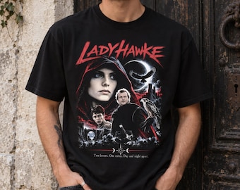 May include: A black short-sleeve t-shirt featuring a graphic print from the movie Ladyhawke. The design includes a hooded figure, two men, a hawk, a crescent moon, and the text "LADYHAWKE" in red, with "Two lovers. One curse. Day and night apart."