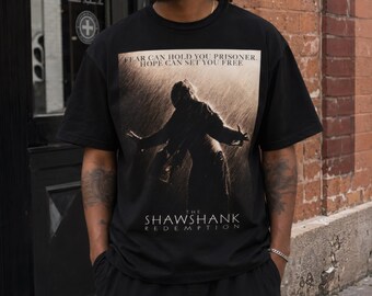 May include: Black t-shirt featuring a movie poster design. The graphic includes a silhouette of a person with outstretched arms in the rain, with the text "FEAR CAN HOLD YOU PRISONER. HOPE CAN SET YOU FREE" and "THE SHAWSHANK REDEMPTION".