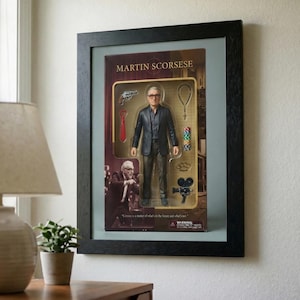 May include: Framed action figure of Martin Scorsese in a black frame. The figure is in a box with accessories like a gun, tie, rosary, and film reel. The box has the name "MARTIN SCORSESE" at the top.