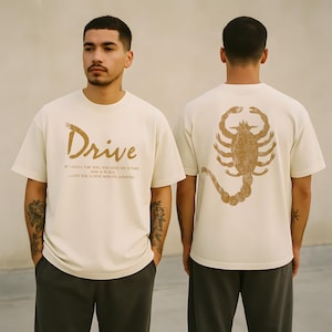 May include: Cream-colored t-shirt with the word "Drive" in a gold script font. Below the word are additional lines of text. The back of the shirt features a large gold scorpion graphic. The shirt is a casual, unisex style.