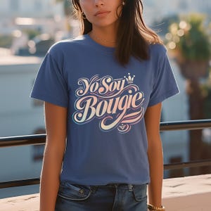May include: A blue t-shirt with the text "Yo Soy Bougie" in a cursive, multi-colored font. The design includes a crown and heart. The shirt is short-sleeved and made of a soft material.