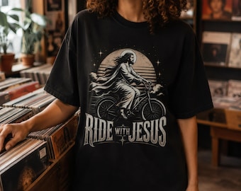 May include: A black short-sleeve t-shirt featuring a graphic illustration of a figure with long hair riding a bicycle in front of a large circular moon. The text RIDE WITH JESUS is printed in a stylized font below the illustration.