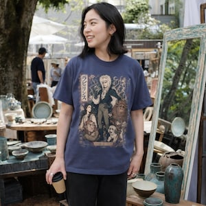 May include: A dark blue short-sleeve t-shirt featuring a graphic print of characters from the film Labyrinth, including Jareth the Goblin King, Sarah, Hoggle, Ludo, and a dog, with Japanese text and floral accents printed on the front.