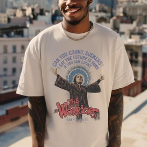 Puede incluir: Camiseta de color crema con un diseño gráfico de la película "The Warriors". El diseño incluye una figura con los brazos extendidos, un halo azul y el texto "CAN YOU COUNT, SUCKERS?" y "THE WARRIORS".