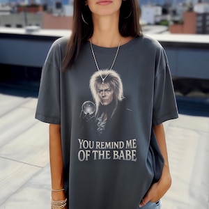 Pode incluir: Camiseta cinza escuro com um retrato de um homem segurando uma bola de cristal. O texto "YOU REMIND ME OF THE BABE" está impresso abaixo. A camiseta tem mangas curtas e gola redonda.