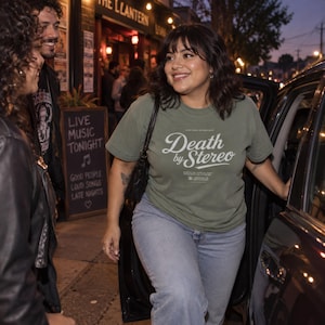 A person enters a car at night outside a bar, wearing an olive green t-shirt with white Death by Stereo text.