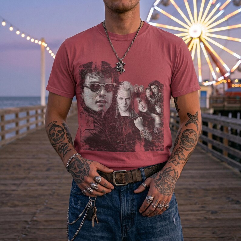 A person with tattoos on their arms stands on a pier, wearing a dark red t-shirt with a distressed graphic of five people, including two men wearing sunglasses.