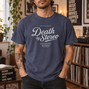 A person with tattooed arms poses in a room with records and posters, wearing a slate blue t-shirt with white Death by Stereo text.