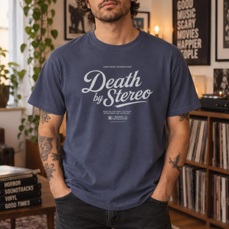 A person with tattooed arms poses in a room with records and posters, wearing a slate blue t-shirt with white Death by Stereo text.