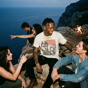 Five people sitting around a campfire on a cliff overlooking the ocean, one person wearing a white t-shirt with a graphic of people.