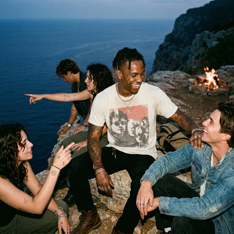 Five people sitting around a campfire on a cliff overlooking the ocean, one person wearing a white t-shirt with a graphic of people.