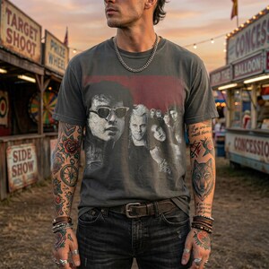 A man with tattooed arms wearing a dark gray t-shirt with a graphic of five people in a red box, standing in front of carnival game booths.
