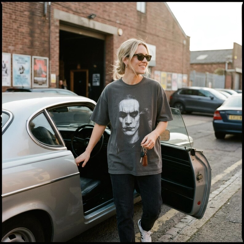 A blonde woman in sunglasses smiles while opening a car door, wearing a charcoal gray t-shirt with a grayscale portrait of Brandon Lee as The Crow.
