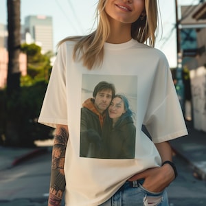 Puede incluir: Una camiseta blanca con una imagen impresa de una pareja. La pareja está de pie uno junto al otro, el hombre lleva una chaqueta marrón y la mujer una chaqueta verde. La imagen tiene un estilo vintage.