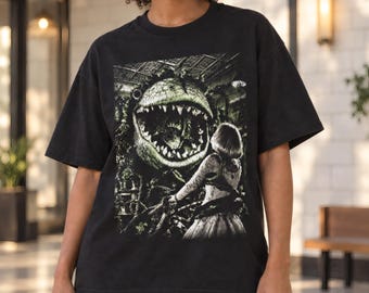 May include: A black short-sleeve t-shirt featuring a graphic print of a large, toothy, green carnivorous plant with a wide-open mouth facing a person in a dress. The detailed illustration includes dark, textured foliage and an indoor setting.