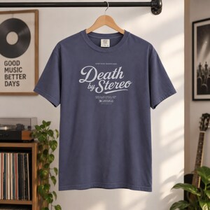 A slate blue t-shirt with white Death by Stereo text hangs on a wooden hanger against a white wall.