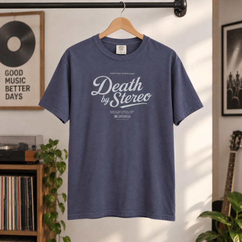 A slate blue t-shirt with white Death by Stereo text hangs on a wooden hanger against a white wall.