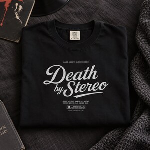 A folded black t-shirt with white Death by Stereo text printed on the chest rests on a dark surface next to a book and blanket.